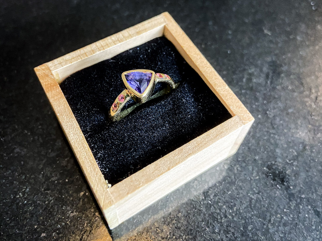 This custom gay/LGBTQ+ engagement/wedding ring features purple sapphire and padparadscha sapphire set in 18K Fairtrade Gold.